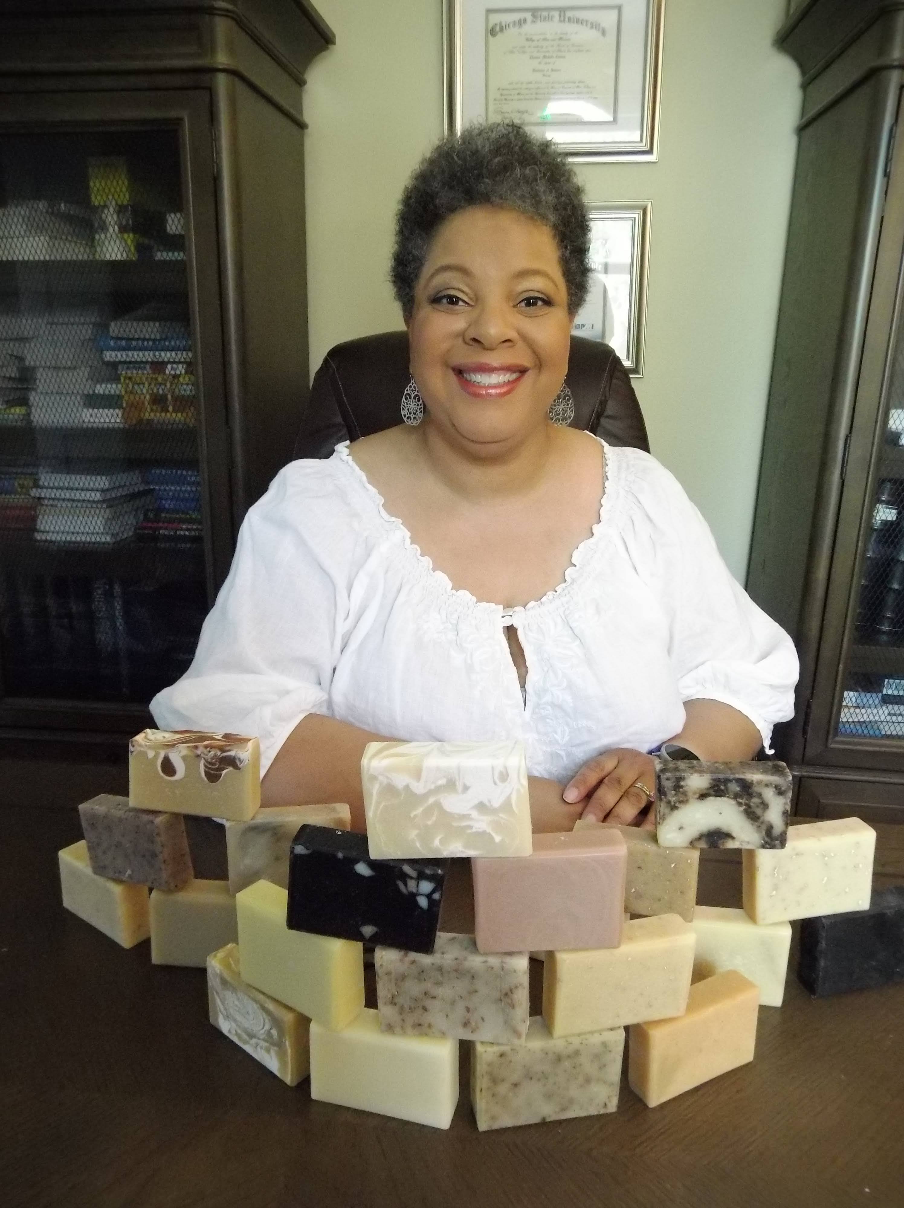 4Elements Bath Founder Charise Cowan-Leroy in a white shirt smiling behind a stacked display of brown, white, and charcoal soaps.