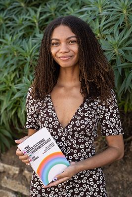 Leah Thomas smiling softly at the camera, holding her book "The Intersectional Environmentalist". She is in a black dress with a daisy pattern standing in front of a foliage background.