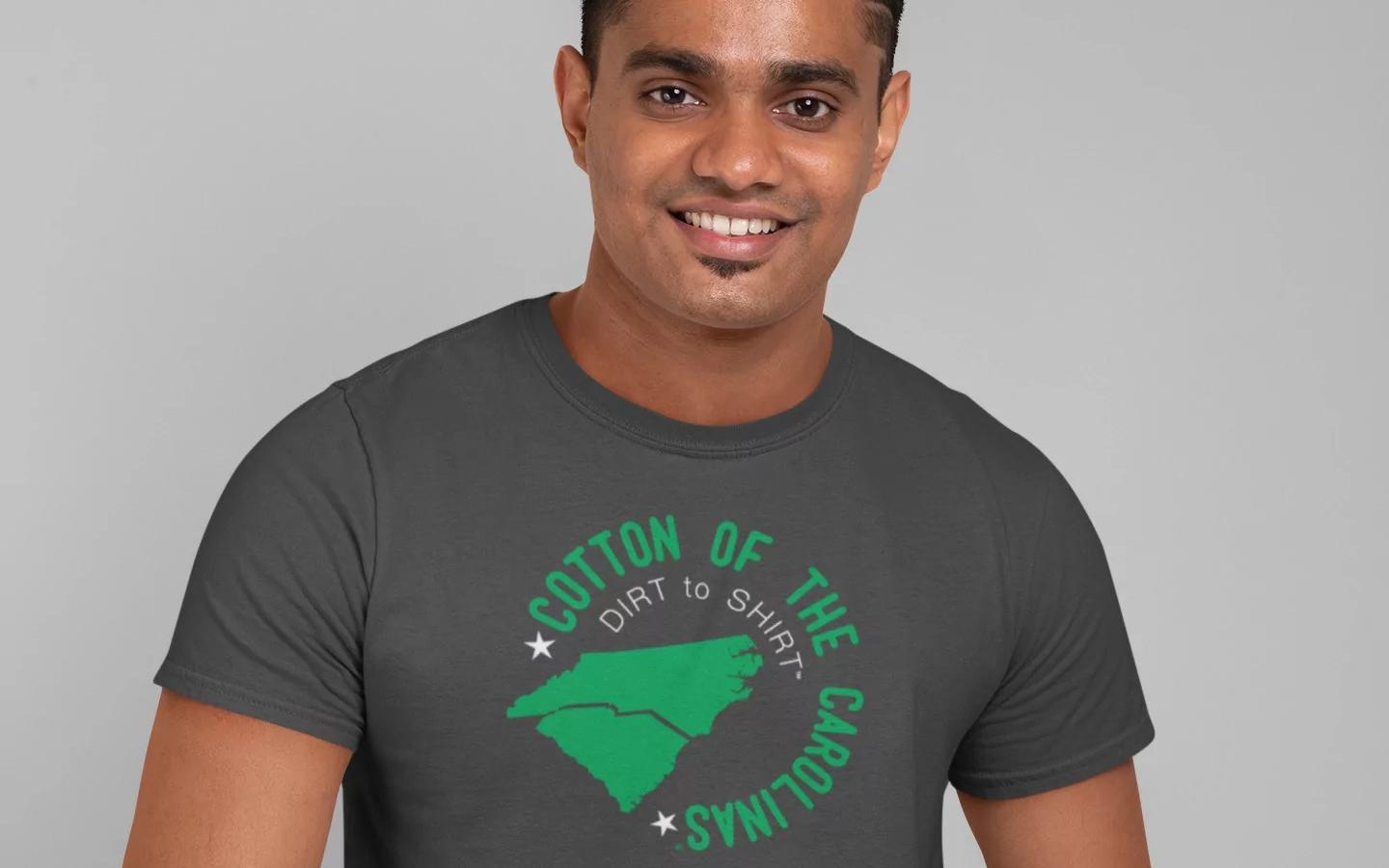 A man of color poses against a grey wall in a dark grey t-shirt made by TS Designs. The shirt reads in green font: "Cotton of the Carolinas. Dirt to Shirt." Small business benefits community.