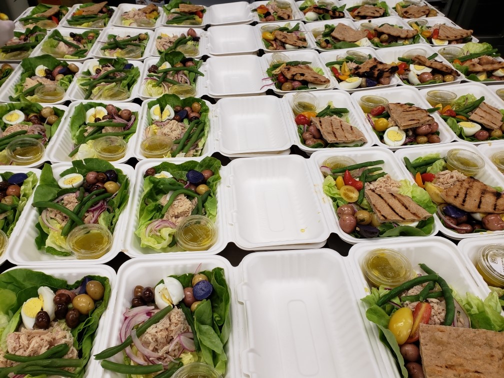 six rows of lunch boxes in biodegradable packaging filled with green veggies, olives, pita, and onions.