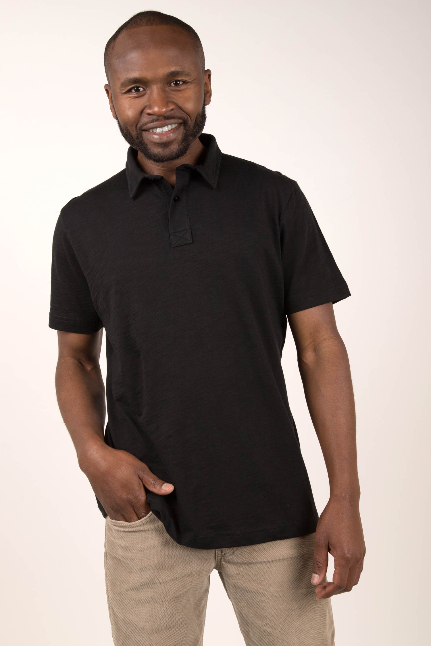 Black male model smiling at camera wearing a Black alpaca wool sub polo shirt. He has one hand in his pocket and is leaning on his right leg.