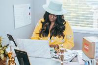 A Black woman sits at a desk in front of a laptop. She's wearing a wide-brimmed white hate and yellow floral blouse, she's looking down at the laptop. On the desk are various items - a picture frame, a box, a statue, a notebook. On the wall next to her is a whiteboard with scribbles on it and behind her is a window. Black-Owned Businesses for Black History Month.