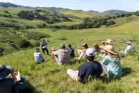 Jackie Eshelman teaching a 5-Day Ecological Outcome Verification™ course at the Center for Regenerative Agriculture at Jalama Canyon Ranch. There are people sitting on the grass all around listening to her speak with rolling green hills in the background.