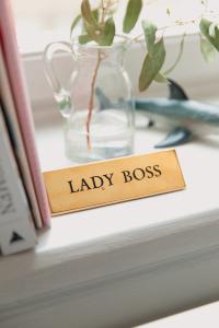 A gold plaque that reads "LADY BOSS" sits on a white shelf. To the left of the plaque are some books, behind the plaque is a plant in a clear, glass jug with a shark figure beside the jug. Women Business Owners.