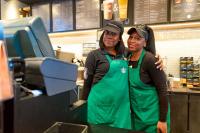 two Black women Starbucks baristas are smiling at the camera. One has her arm wrapped another the other in a side hug. They look prepared and ready for work.