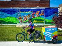 A woman is riding a green bike with a box attached to the back that reads "Topeka & Shawnee County Public Library." She is in front of a mural that says "Greetings from Topeka, Kansas."