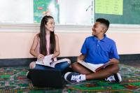 An elementary school aged girl and boy sit on the floor with books in their laps. They are smiling at each other.