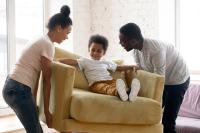 A Black mom and dad pick up an armchair with a 4 year old kid sitting in it. They're all smiling and laughing together.