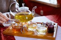 A wooden tray with tea in a glass kettle, jars of jam, and an open mason jar of honey. A hand sticks out from the left holding a honey dipper. Sustainable honey products