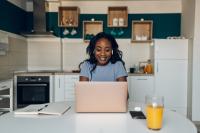 A young Black woman looks for financial information on her laptop in her kitchen
