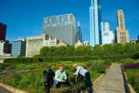 Two adults, a white man and white woman, kneel in a garden with the Chicago skyline behind them. A video camera is set up recording them.