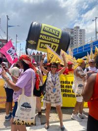 A woman in a patterened white dress stands amidst climate protesters with her arms raised victoriously.