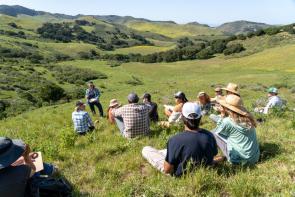 Jackie Eshelman teaching a 5-Day Ecological Outcome Verification™ course at the Center for Regenerative Agriculture at Jalama Canyon Ranch. There are people sitting on the grass all around listening to her speak with rolling green hills in the background.