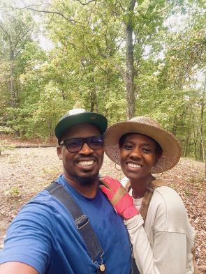 Osei Doyle and Brendalyn King are dressed in overalls and t-shirts, wearing brimmed hats. They are taking a selfie outside, with Brendalyn holding onto Osei's shoulder.