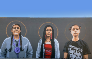 Mural painted on the outside of Native American Bank. There are three women of different ages standing side by side with halos around their heads to represent what Native American Bank honors and celebrates.