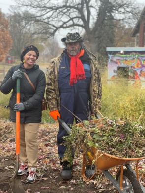 Michael Chaney is wearing a big brown coat and a red scarf, holding a wheelbarrow filled with foliage for compost. He stands next to Jasmira Colon who is smiling at the camera and holding a shovel with an orange handle.
