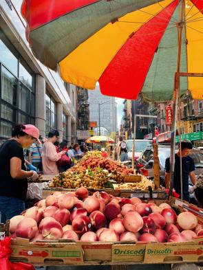 a fruit market in NYC. Close up is a stand of nectarines under a red, yellow, and blue sun parasol. In the background are more fruit stands and onlookers.