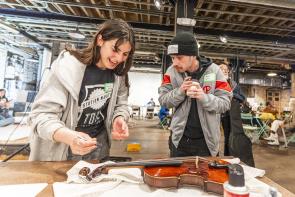 A woman with a staff shirt is explaining how to repair and clean a violin. A man holding a coffee cup is watching her work.