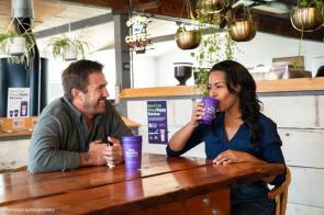 Two people, a white man and a mixed descent woman, sit at a coffee table in a cafe. He is looking at her and smiling. She is taking a sip from a reusable purple cup.