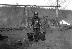 A black and white photo of a child under the age of 12 carrying two grease pails. He is covered in grease on his body. He stands alone in a bare and gray construction site on a hill.