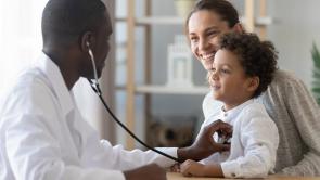 A young black doctor holds a stethoscope up to a mixed child's heart. The mom has the child on her lap. Both are smiling at the doctor.