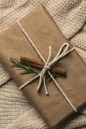 "Gift wrapped in brown kraft paper tied with natural cotton twine and decorated with cinnamon sticks and fresh rosemary sprigs on a knitted blanket.
