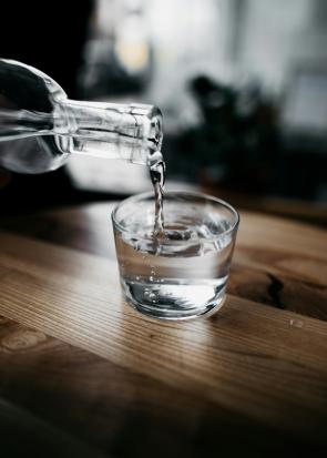 Water pouring from a glass bottle into a glass jar, which is set on a wooden table.