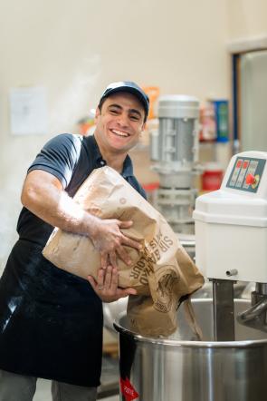 A small business owner pours a bag of flour into a bowl in his bakery