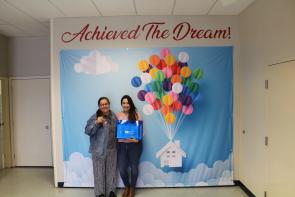 A woman and her daughter stand in front of a sign that reads, "Achieved the Dream" after they became homeowners.