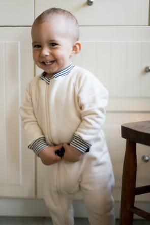 Baby standing in front of kitchen cabinets wearing a natural cotton fleece footie that has a collar and cuffs made from grey and natural pin stripe fabric. 