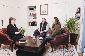 Athens Impact Socially Responsible Investments' founder Michelle Williams sits in a brown leather armchair - on her right are two clients, a white man in a black vest and a white woman with long brown hair in a green sweater, sitting in brown armchairs. Michelle has short hair, wears a black pantsuit, and has a folder in her hands.