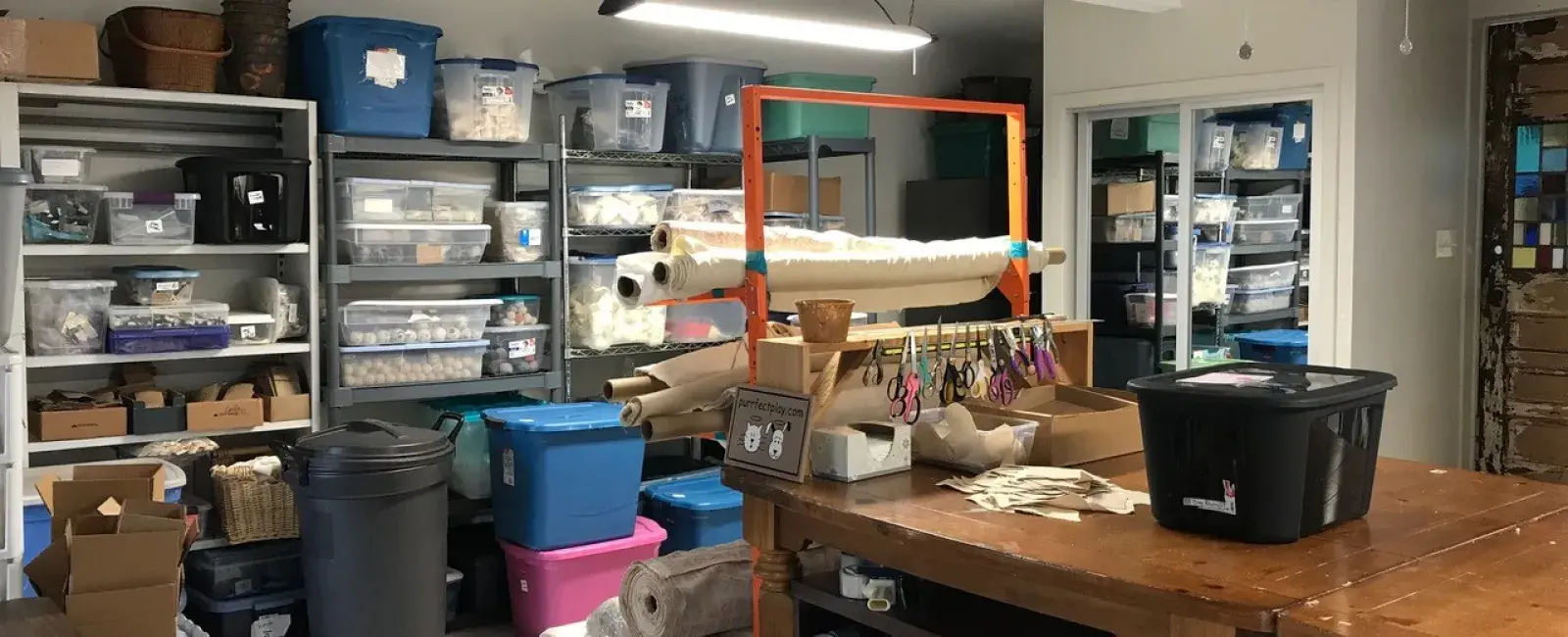 A craft studio, featuring a big wooden table in the middle with a black, plastic bin on top. Against the wall in the back are shelves with lots of bins and craft materials. Woman Business Owners.