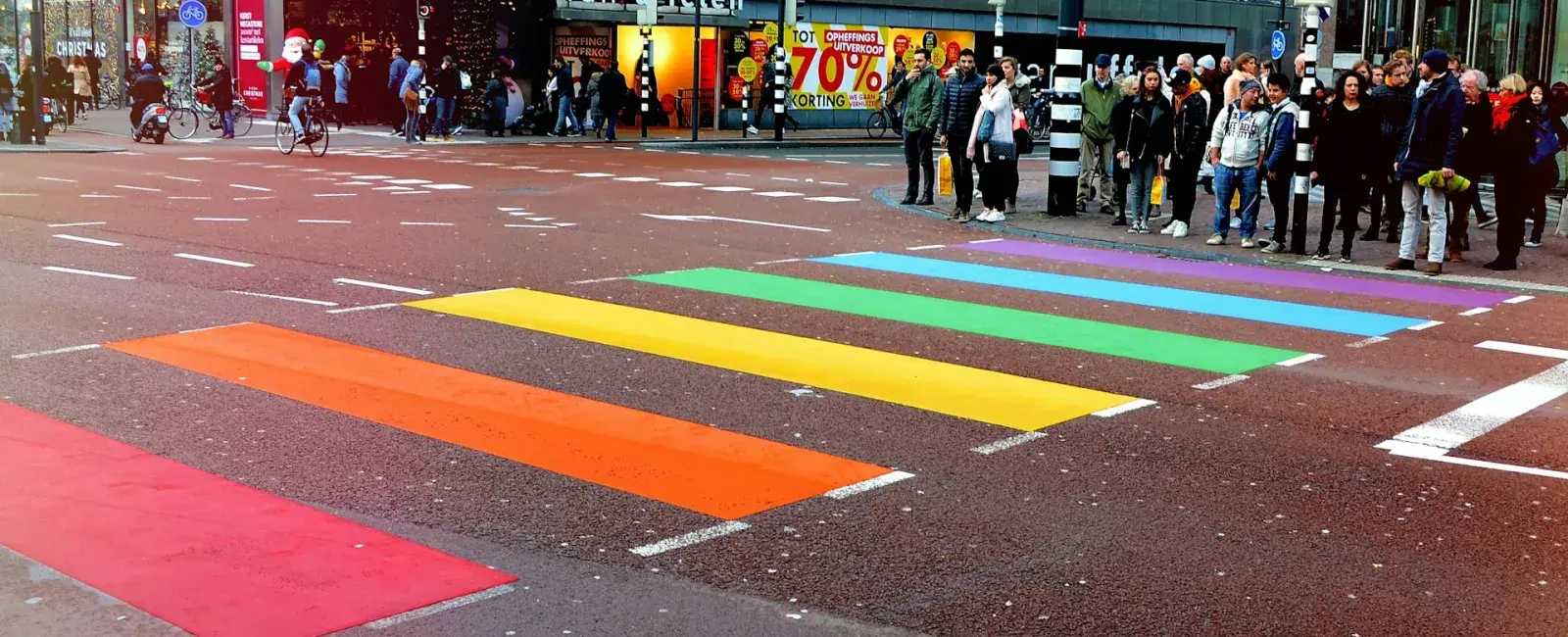 A rainbow crosswalk at an intersection with people waiting to cross. LGBTQ-owned