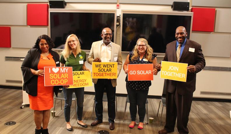 Image: people stand holding signs that say "I love my solar co-op, "Solar = Energy Freedom," "We Want Solar Together!," "Go Solar," and "Join Together." Title: Getting to 100% Clean Energy, Equitably