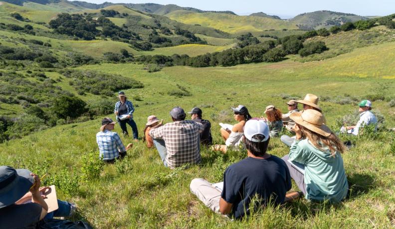 Jackie Eshelman teaching a 5-Day Ecological Outcome Verification™ course at the Center for Regenerative Agriculture at Jalama Canyon Ranch. There are people sitting on the grass all around listening to her speak with rolling green hills in the background.
