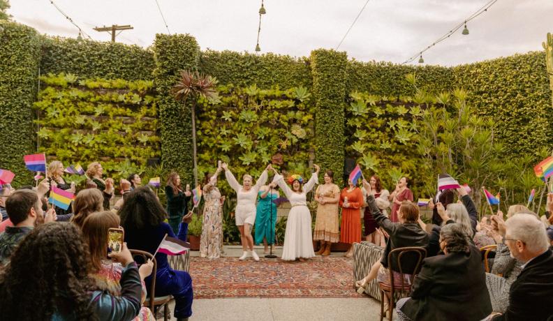 Dana and Anya raise their arms triumphantly during the wedding ceremony. They are surrounded by loved ones waving pride flags and a green, leafy background.