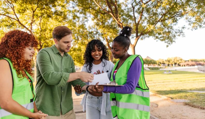 a group of four young people wearing construction vests and button down shirts, looking at a clipboard together. They all have curious and engaging expressions. They are standing outside and its sunny and green.