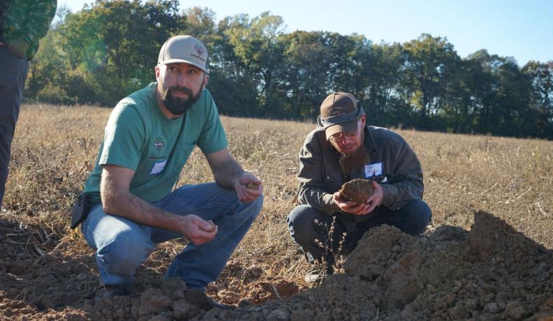 two men squatting in the dirt of a field, holding clumps of soil in their hands and examining them.
