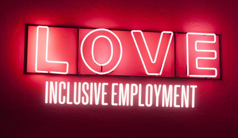 White neon sign against red wall, reading: LOVE INCLUSIVE EMPLOYMENT