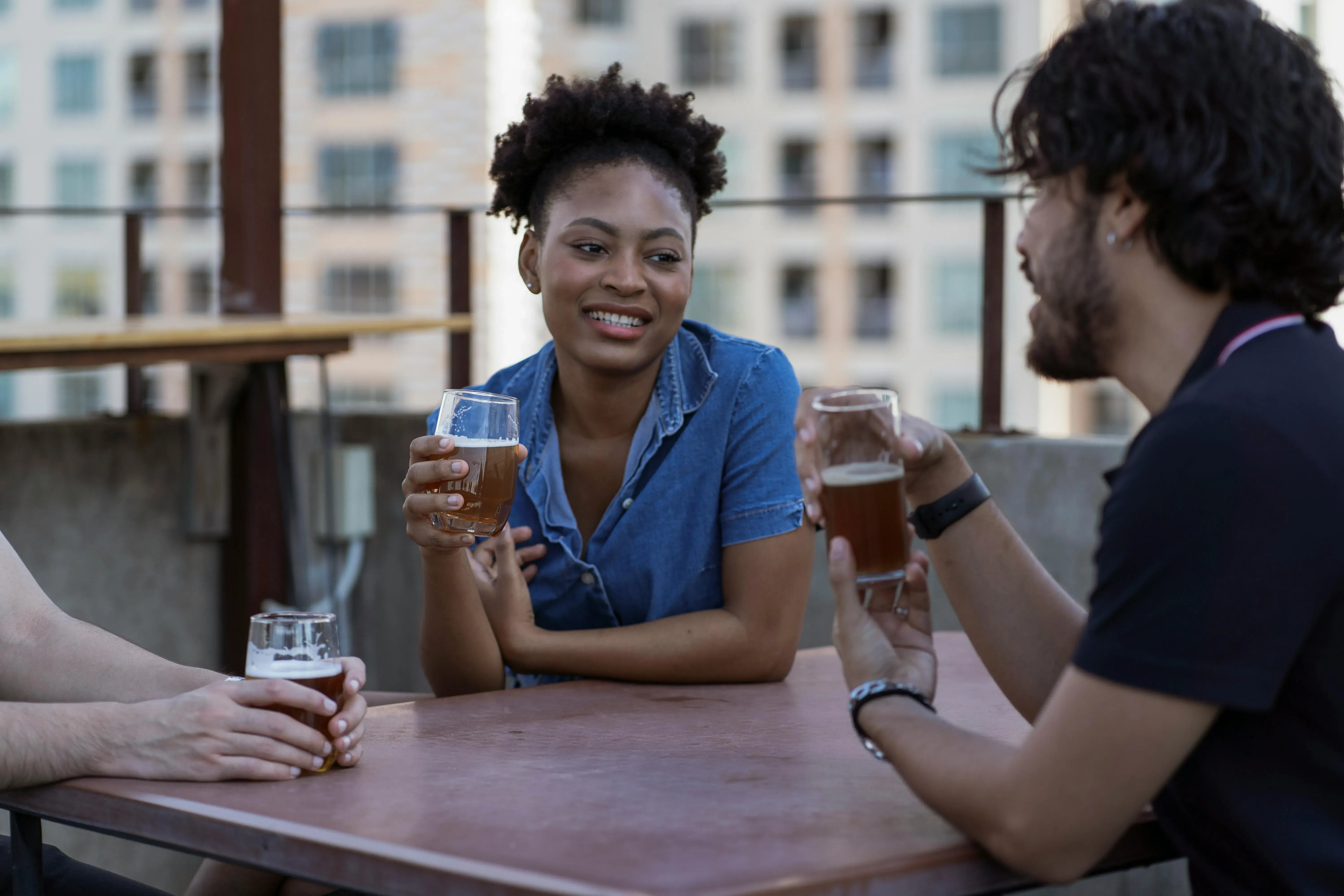 Woman smiling while holding a beer glass at a rooftop bar with two friends, urban buildings visible in the background.