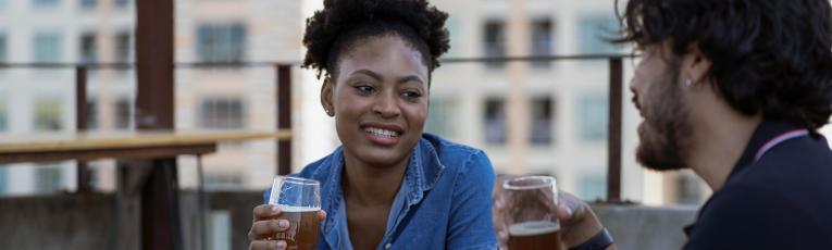 Woman smiling while holding a beer glass at a rooftop bar with two friends, urban buildings visible in the background.