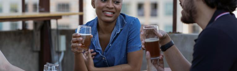 Woman smiling while holding a beer glass at a rooftop bar with two friends, urban buildings visible in the background.
