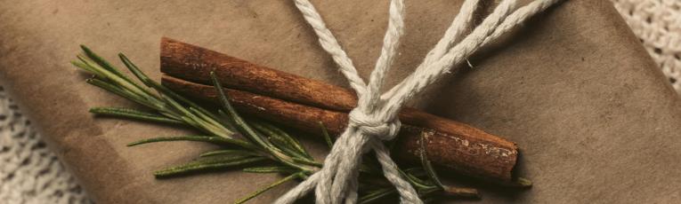 "Gift wrapped in brown kraft paper tied with natural cotton twine and decorated with cinnamon sticks and fresh rosemary sprigs on a knitted blanket.