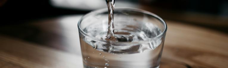 Water pouring from a glass bottle into a glass jar, which is set on a wooden table.