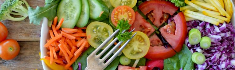 A bowl of salad on a wooden table. The salald contains tomatoes, purple cucumbers, yellow bell peppers, and lettuce, among other things.