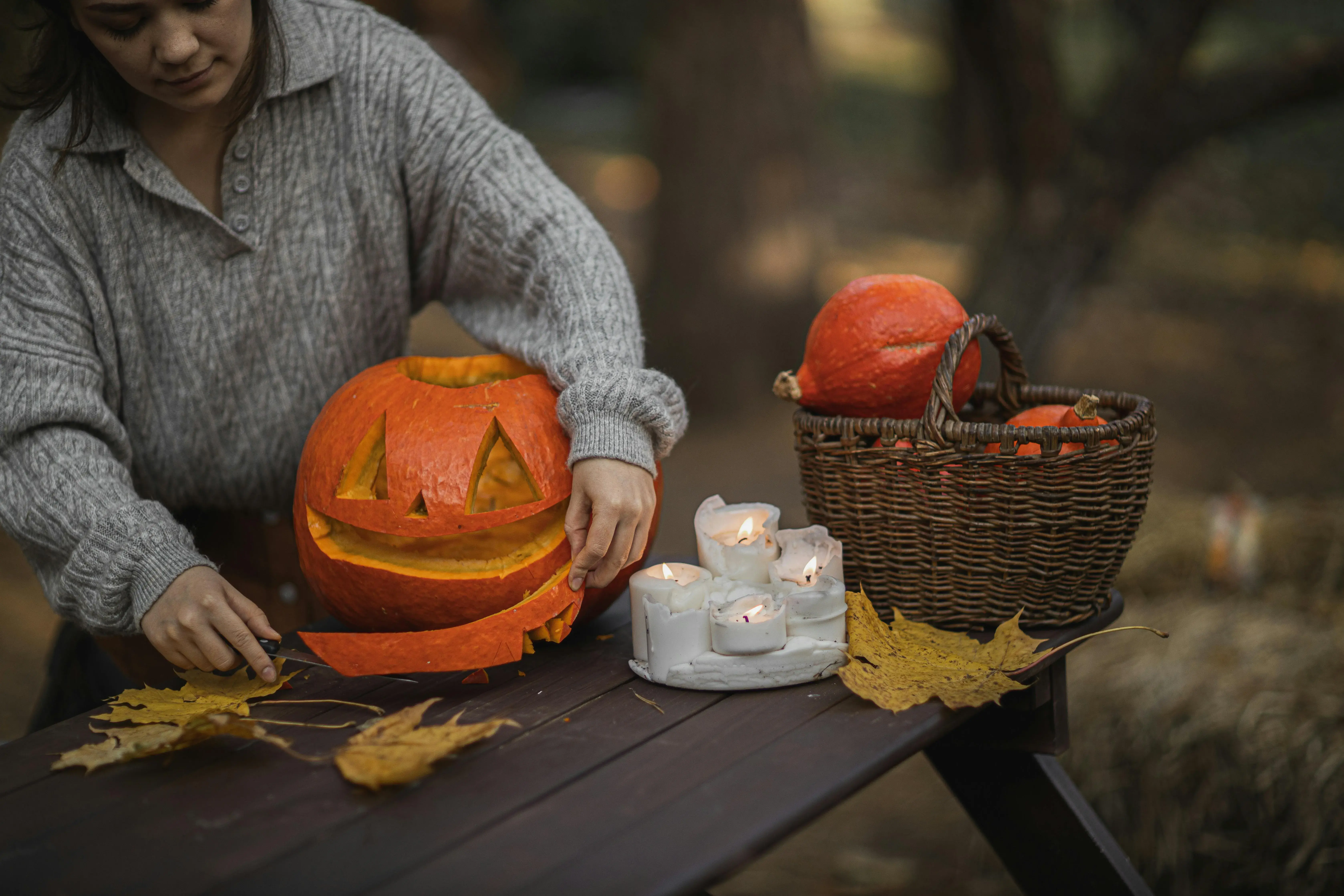 Woman in a gray sweater carving a jack-o-lantern pumpkin at an outdoor table with lit candles and a basket of pumpkins