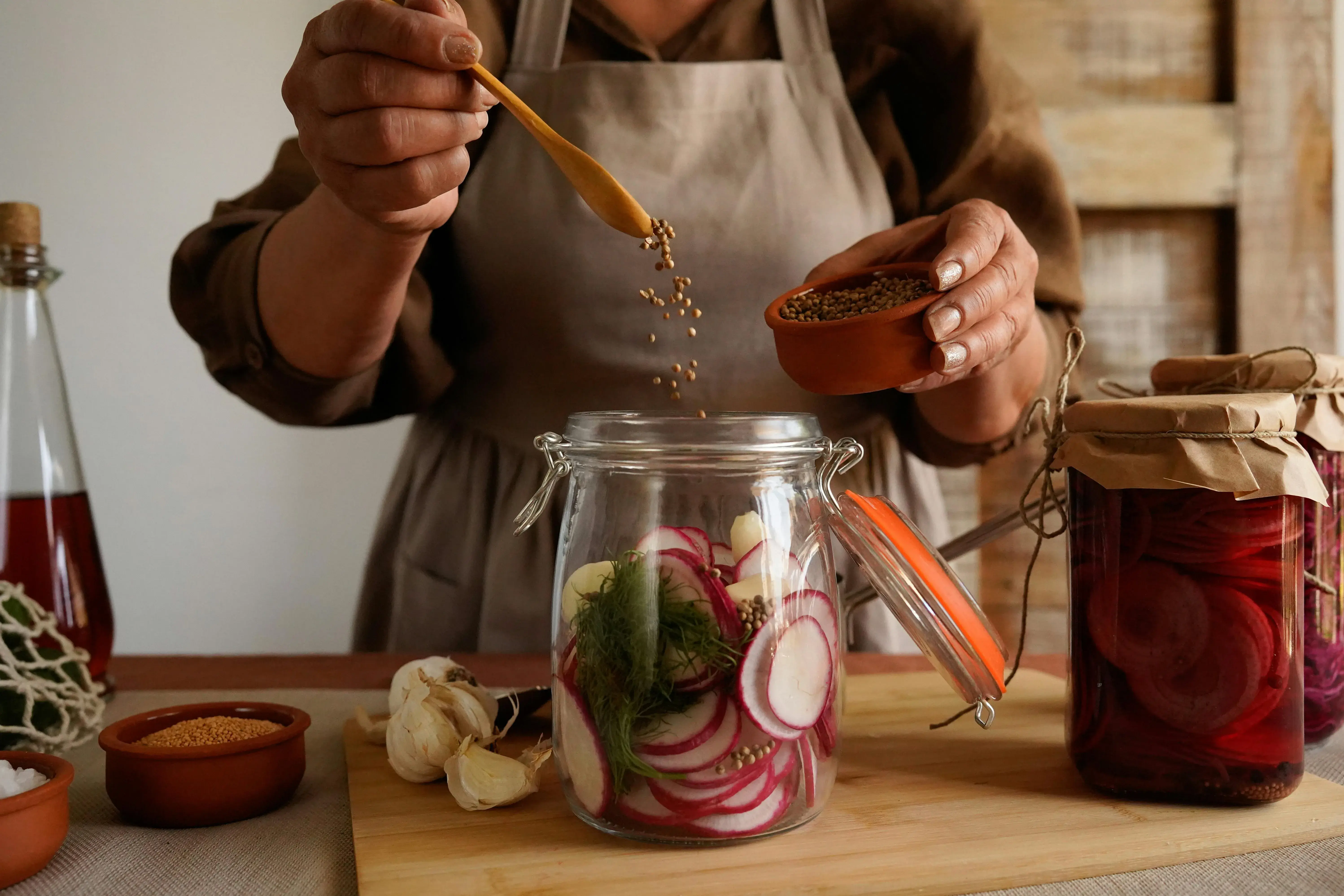 Person in an apron sprinkling seeds from a wooden spoon into a glass jar containing sliced radishes and herbs for pickling