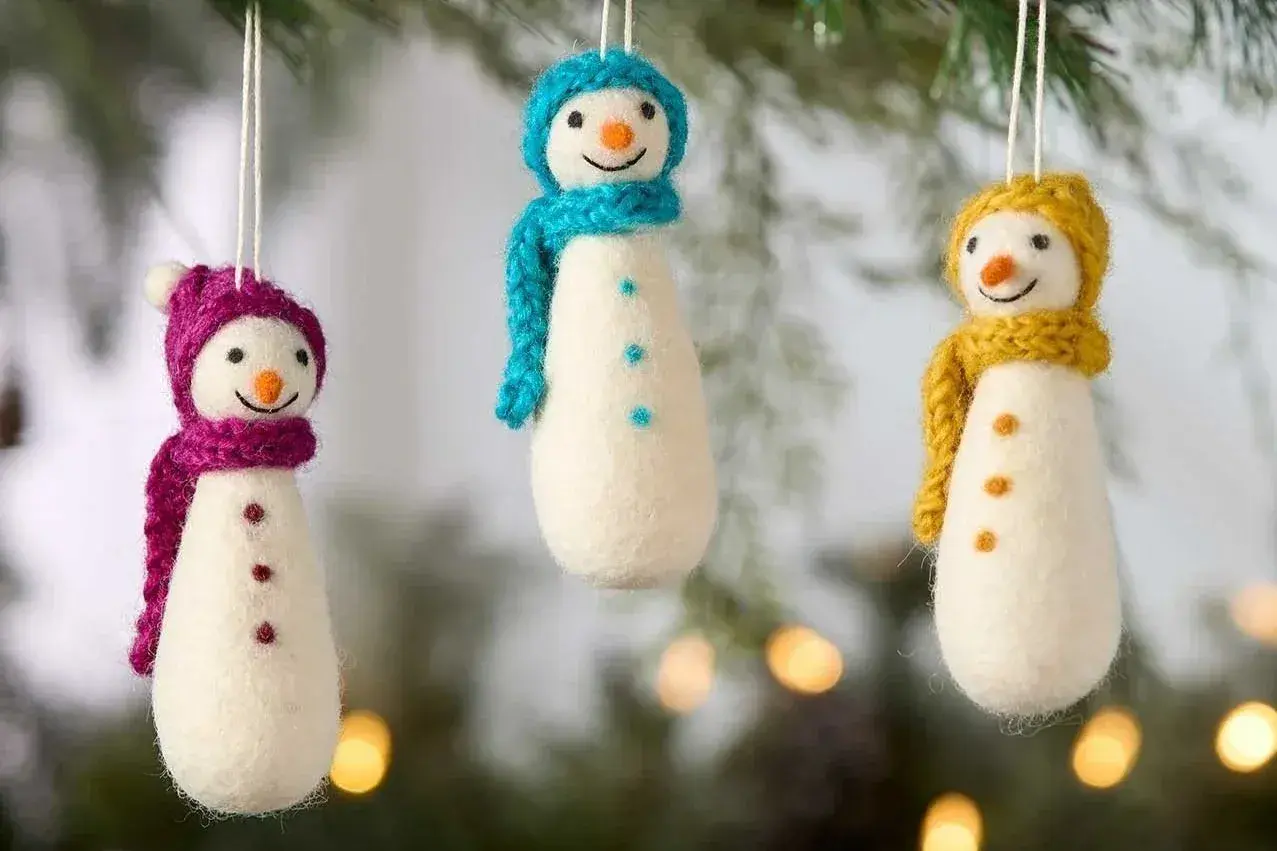 Three handmade felt snowman ornaments with colorful knit scarves in magenta, turquoise, and yellow hanging against evergreen branches.