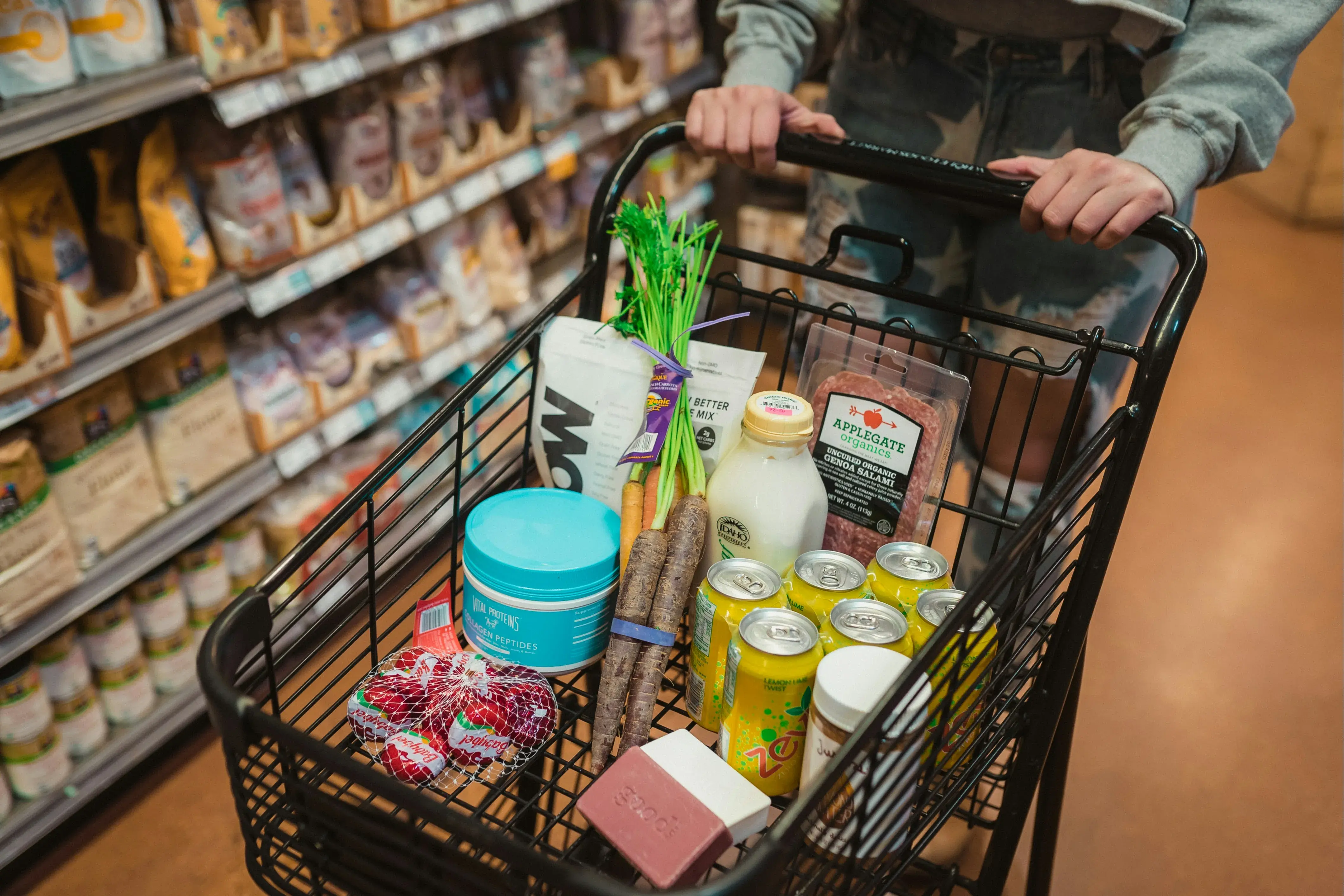 Shopper pushing metal basket filled with groceries including fresh vegetables, packaged foods, and beverages in grocery store aisle.