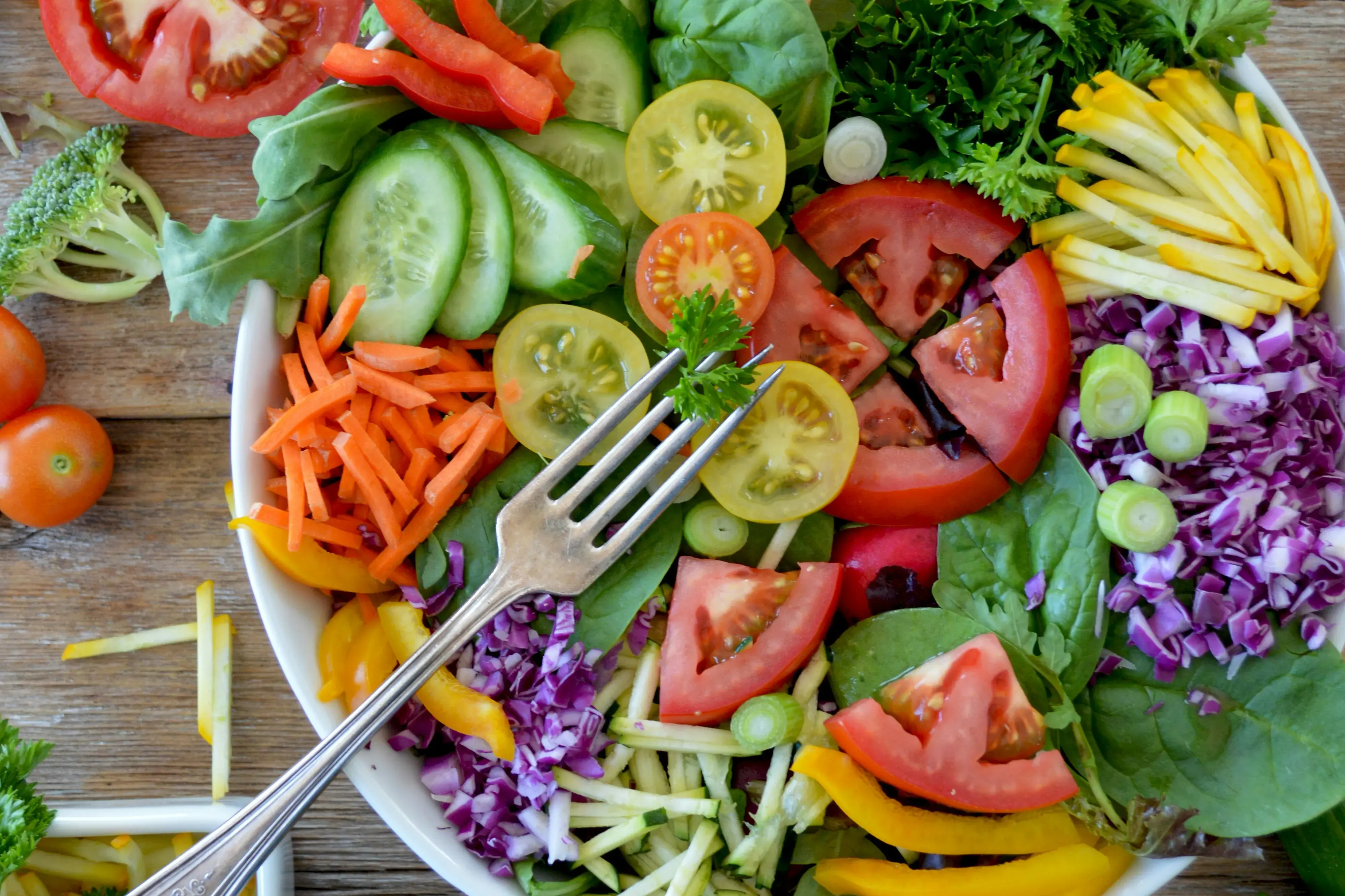 A bowl of salad on a wooden table. The salald contains tomatoes, purple cucumbers, yellow bell peppers, and lettuce, among other things.